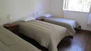 A room with three beds, each with a pillow and a bedside table with a lamp. There is a window with curtains, and a small plant on the wall. - Casa Provence (Santiago)