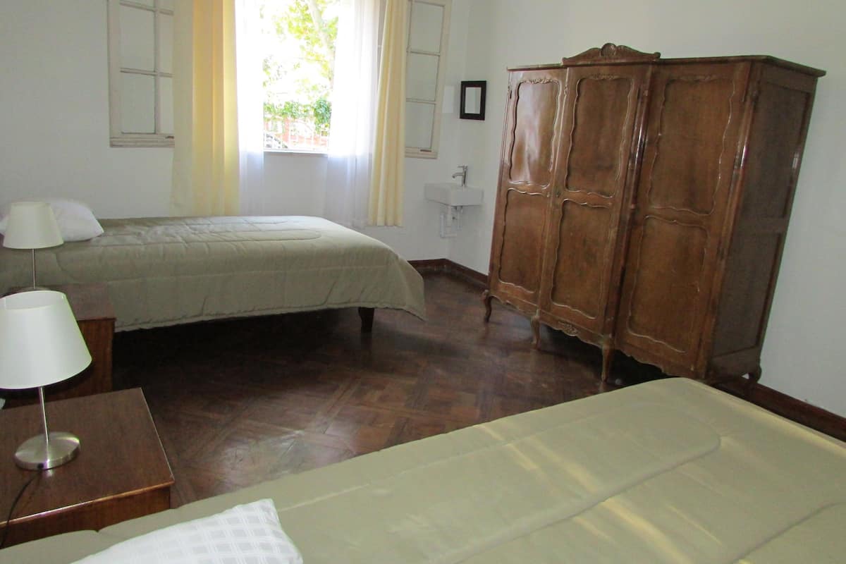 A room with two beds, a wooden wardrobe, a bedside table with a lamp, and a window with curtains.