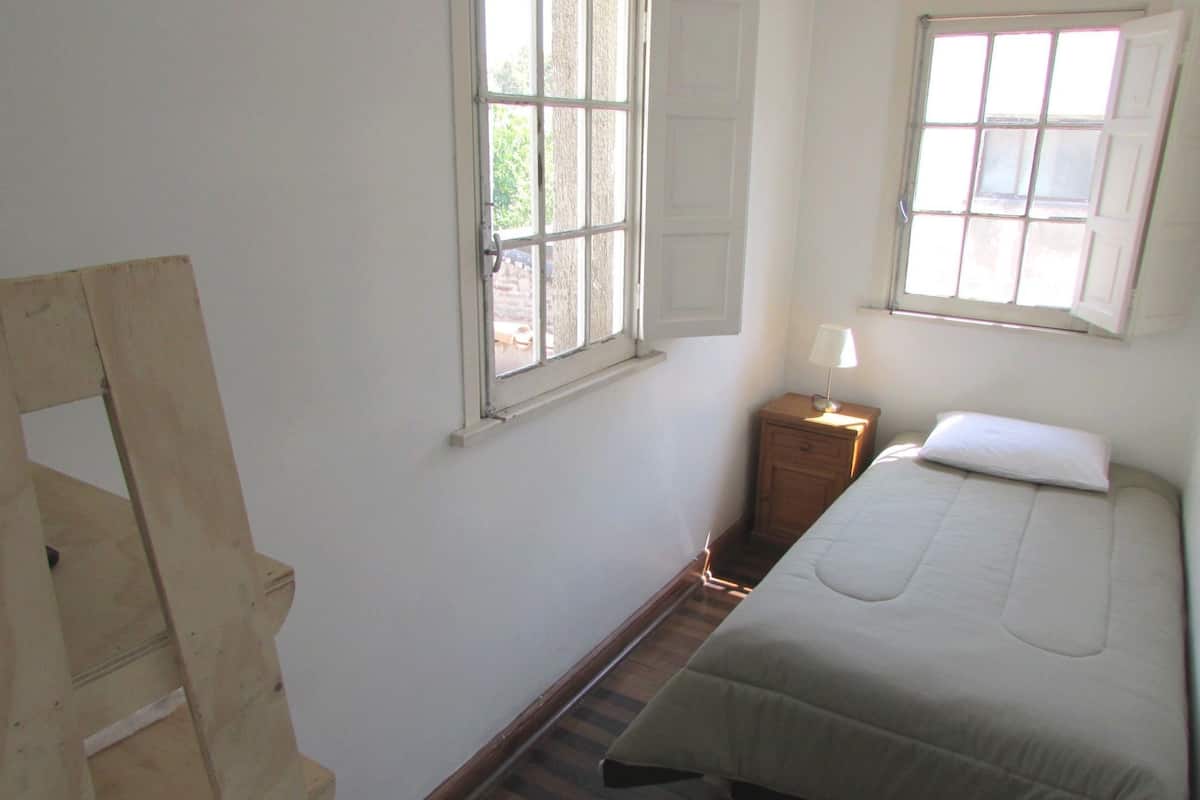 A bedroom with a wooden ladder, a bed with a white mattress and pillow, a nightstand with a lamp, and two windows with shutters.
