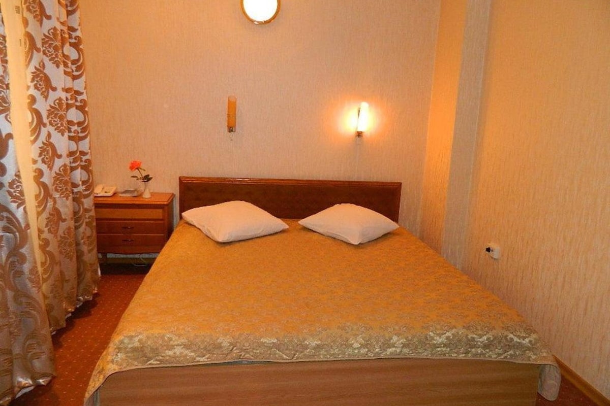 A single bed with two pillows, a bedside table with a lamp, and a curtain with a floral pattern.