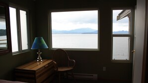 Une chambre à coucher avec un lit, un bureau avec une lampe, une chaise et une vue sur un lac, avec des montagnes en arrière-plan.