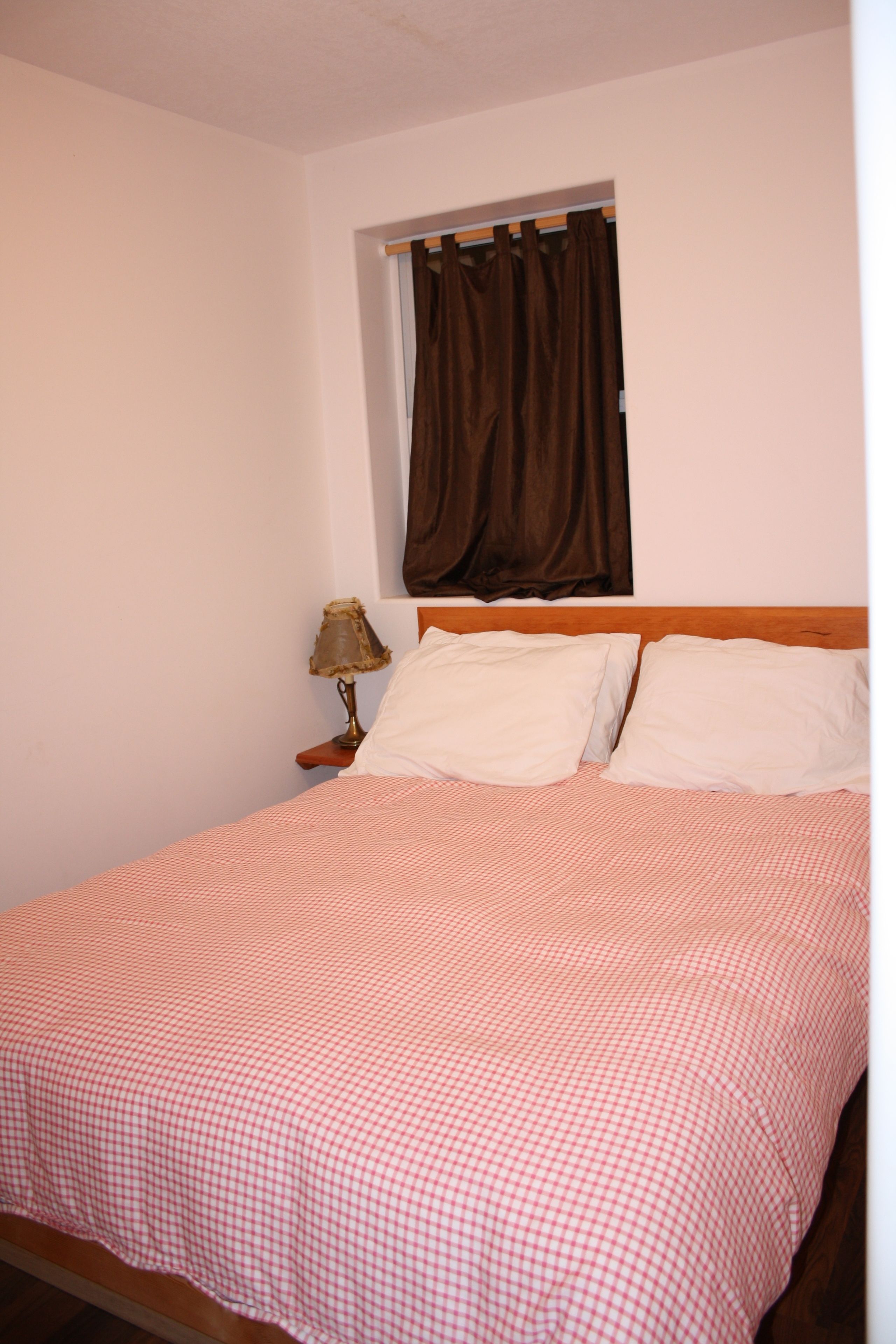 Una cama bien hecha con una colcha a cuadros, un cabecero de madera, una lámpara de noche y una ventana con cortinas marrones.