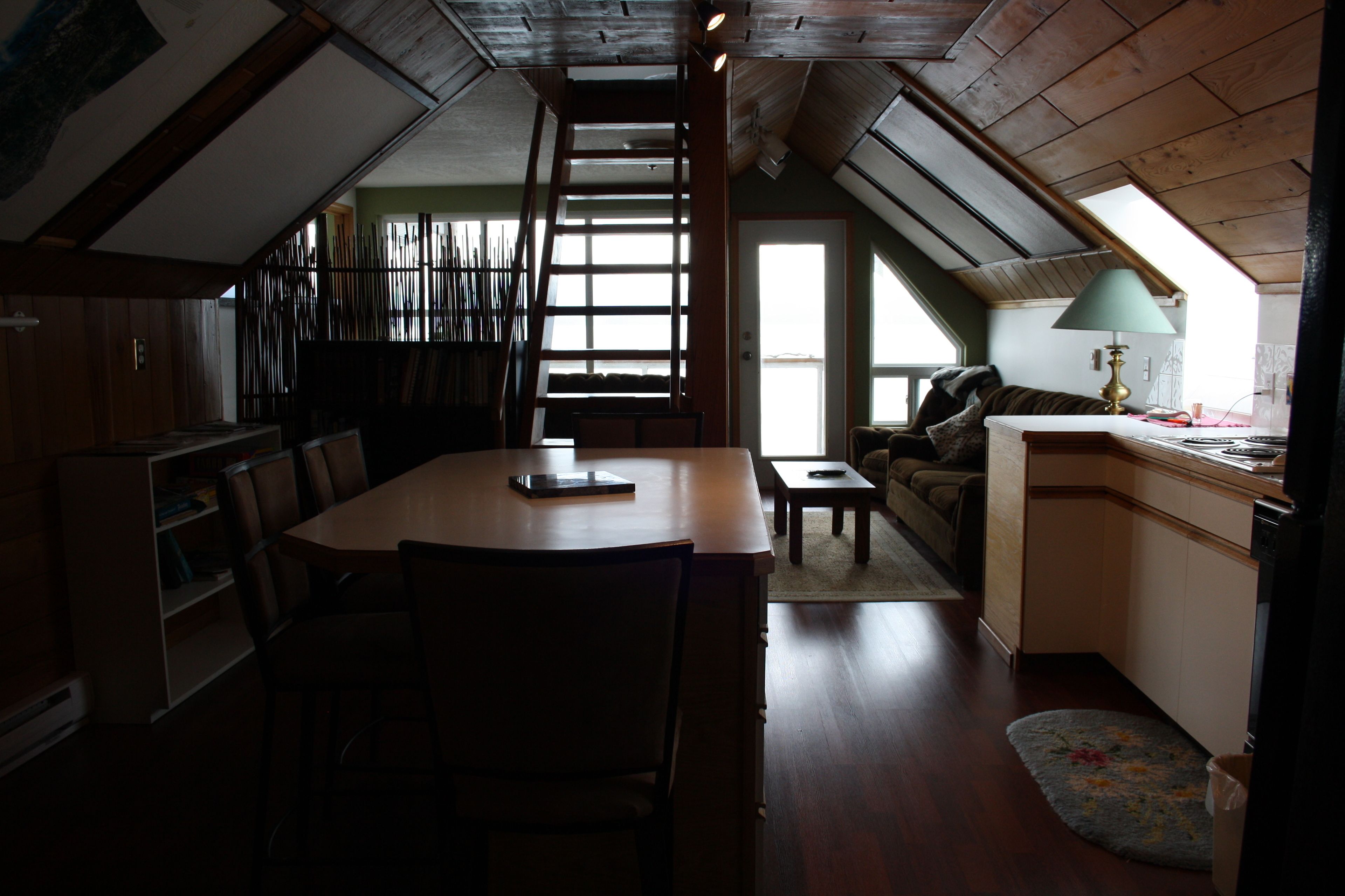 Un acogedor cuarto ático con una mesa de comedor de madera, sillas, una escalera, un sofá y una pequeña cocina con una lámpara.