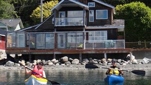 Deux personnes pratiquent le kayak sur une étendue d’eau calme, avec une grande maison en bois en arrière-plan.