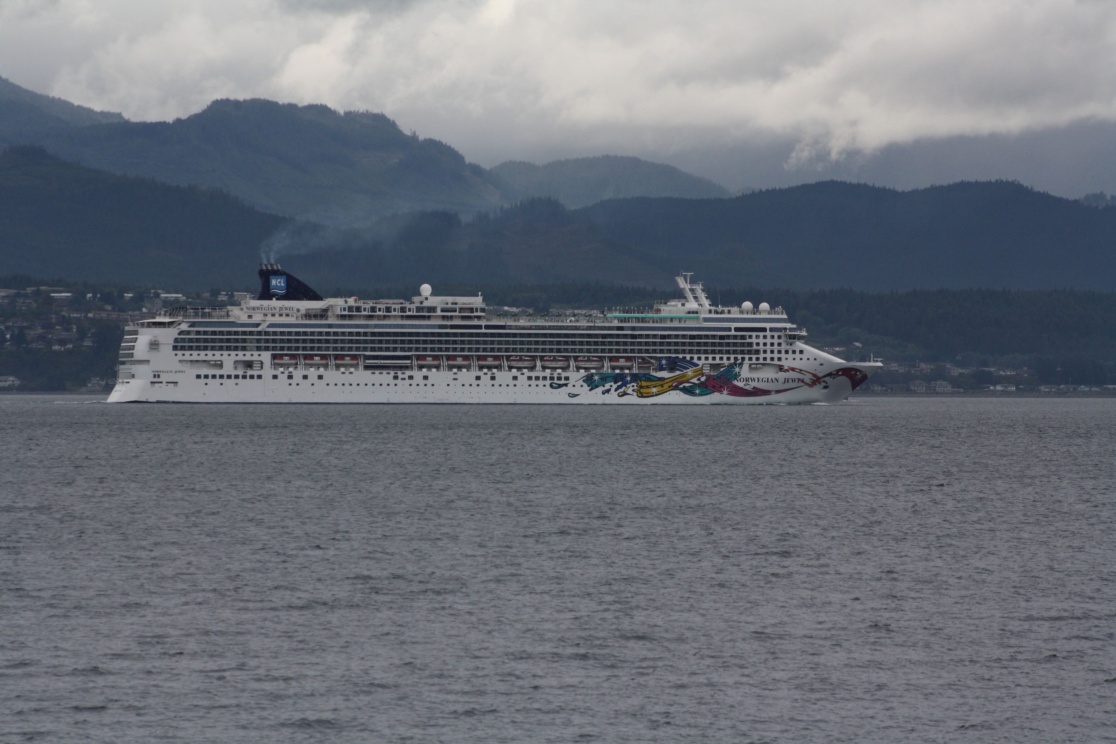 Un grand navire de croisière, doté de nombreux ponts et arborant le pavillon norvégien, navigue sur une étendue d’eau dont le paysage est dominé par des montagnes.
