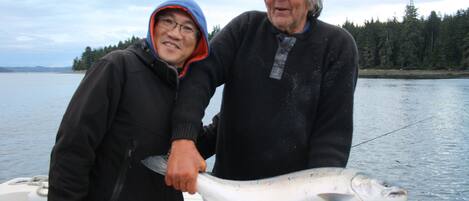 To personer holder en fisk på en båd med en skovklædt kystlinje i baggrunden.