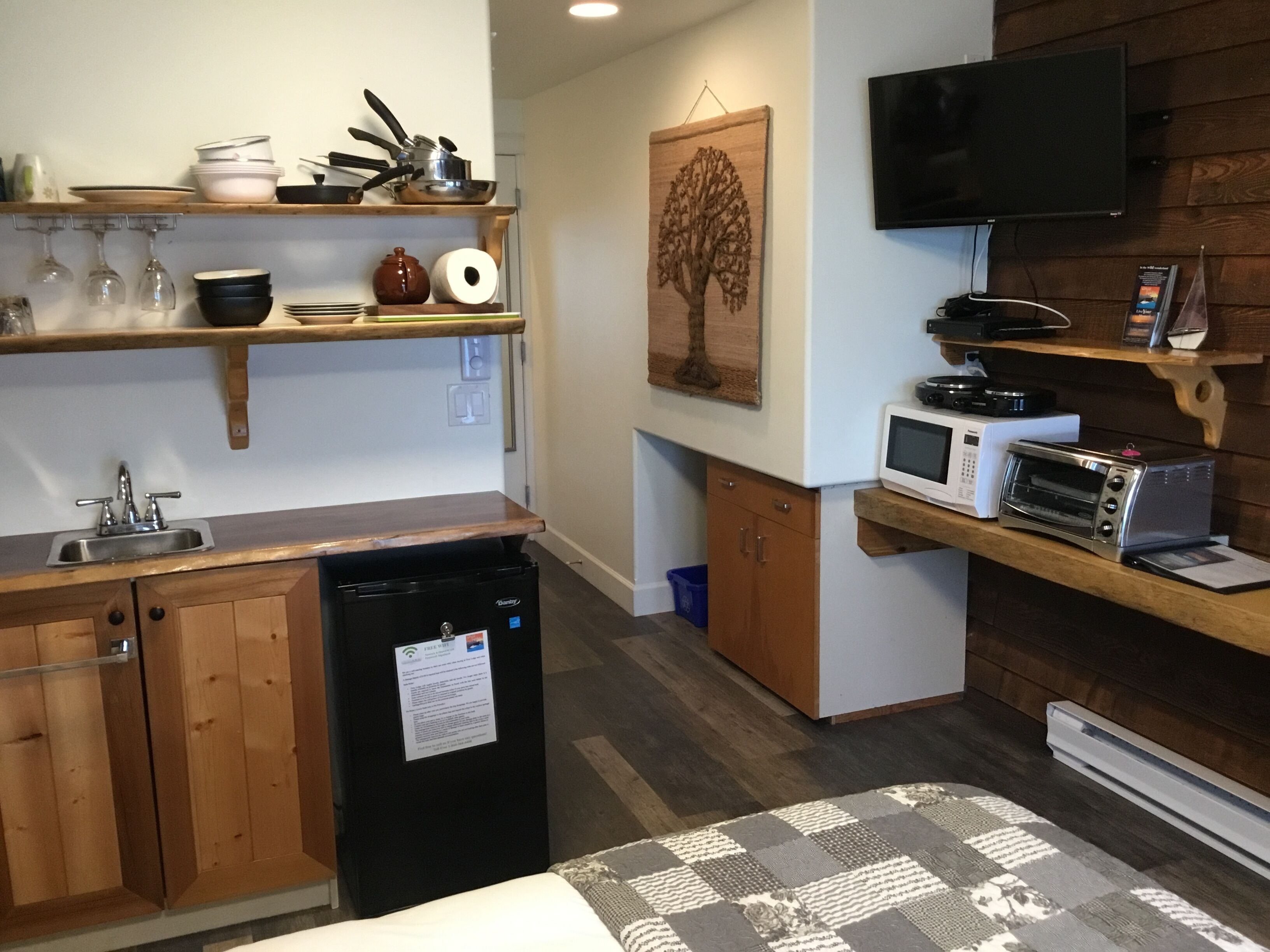 A compact kitchen with wooden cabinets, a built-in sink, a refrigerator, and a microwave. There is a flat-screen TV mounted on the wall and a small shelf with kitchen utensils.