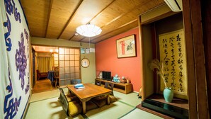 A traditional Japanese room with tatami flooring, a low table, chairs, a television, and a vase with dried plants. - Kumano House (Kyoto)