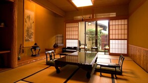 Una habitación con esteras de tatami, una mesa baja y sillas, puertas correderas y una pintura tradicional japonesa.
