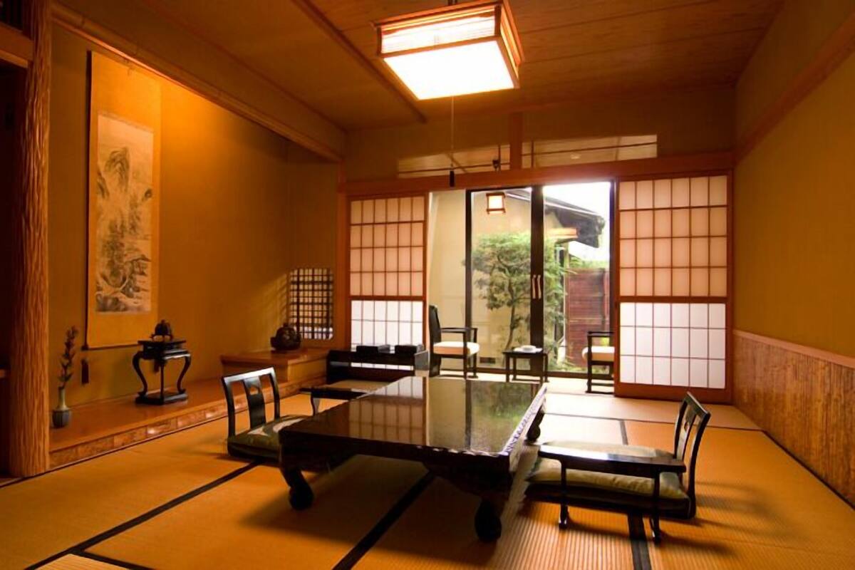 Um espaço com tatames, uma mesa baixa, cadeiras, portas deslizantes e uma pintura tradicional japonesa.