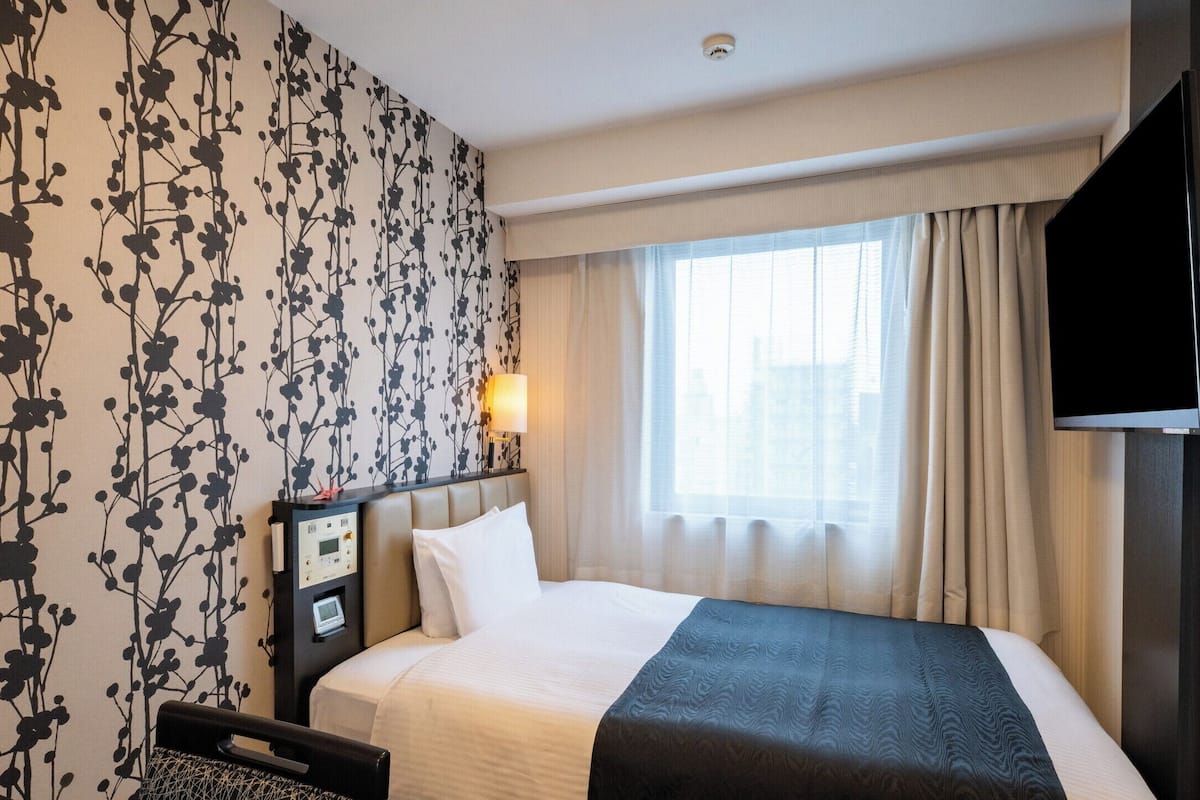 Een hotelkamer met een bed, nachtkastje met lamp, televisie en raam met gordijnen.
