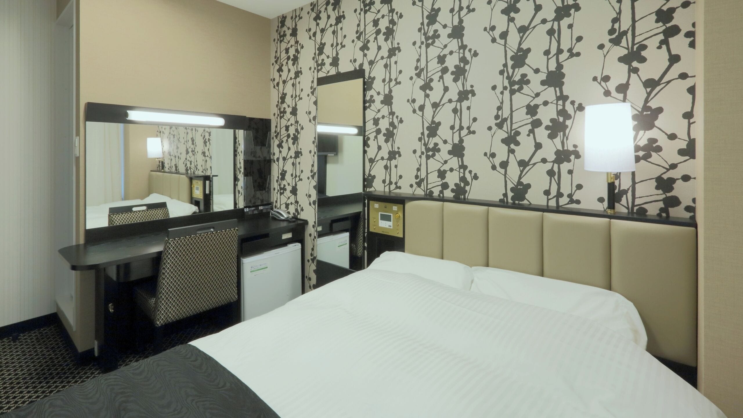Ein Hotelzimmer mit einem Bett, einem Schreibtisch mit Computer, einem Spiegel und einer Wand mit floralem Muster.