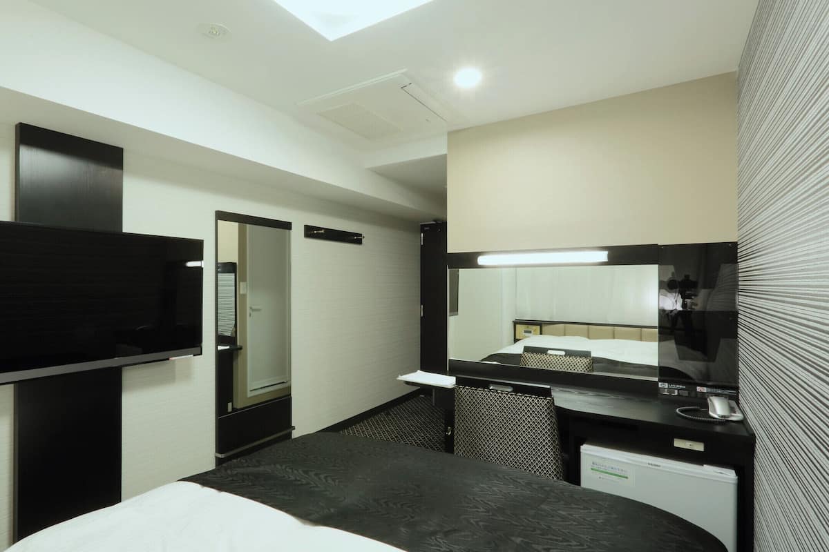 Habitación de hotel con televisor de pantalla plana, espejo grande, mesita de noche, cama y secador de pelo incorporado.