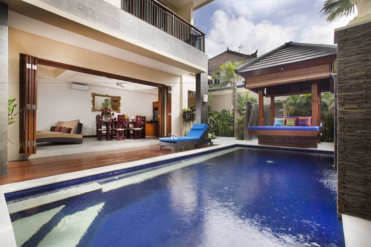 A modern outdoor pool area with a lounge chair, a wooden deck, and a covered seating area.