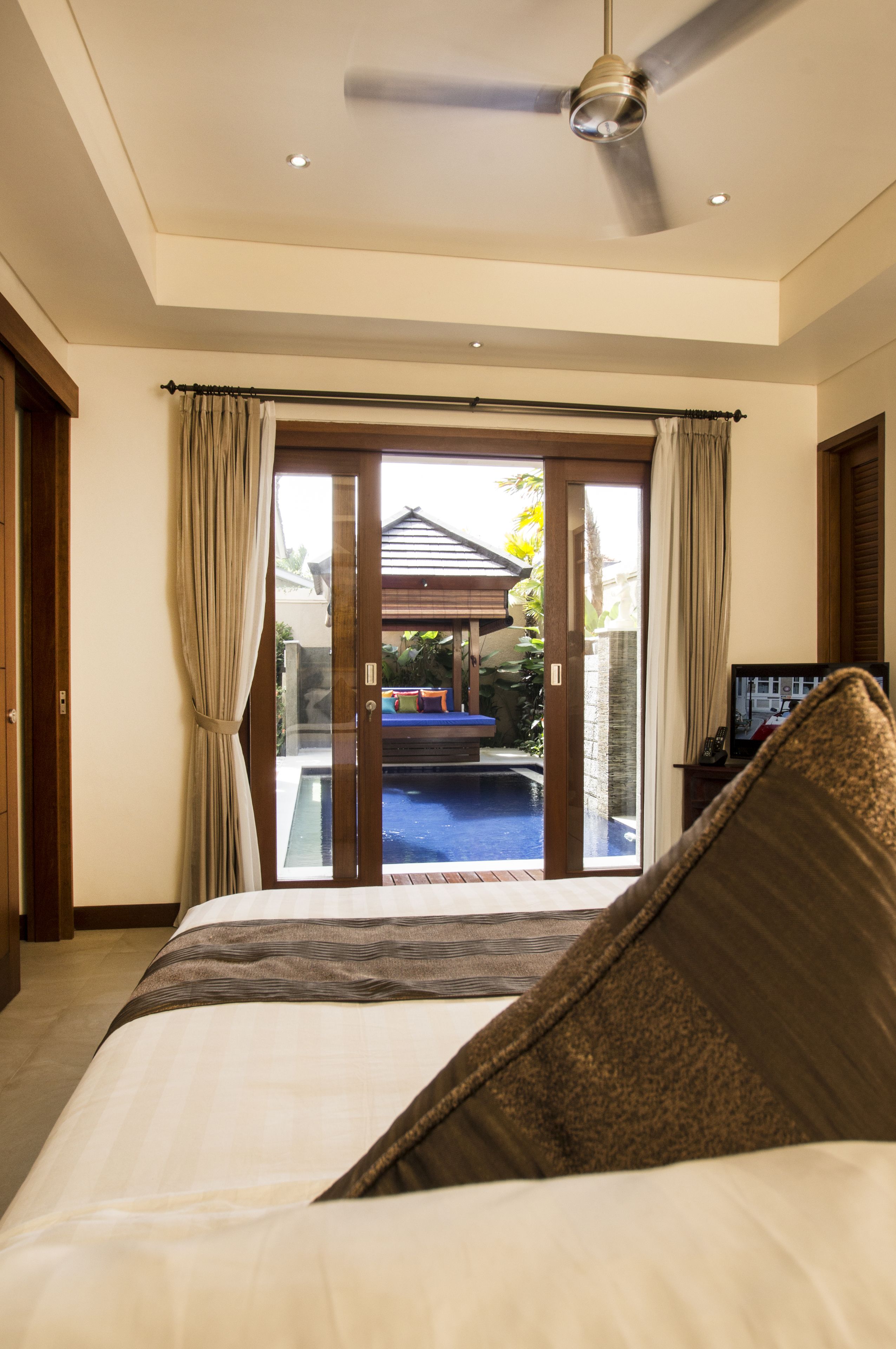 A bedroom with a bed, a ceiling fan, a view of a swimming pool, and a door leading outside.