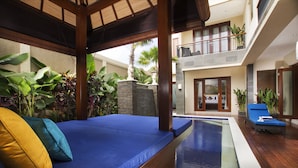 A poolside area with a wooden deck, a blue cushioned seating area, and a view of a building with a balcony. - The Oasis Retreat Seminyak (Seminyak)