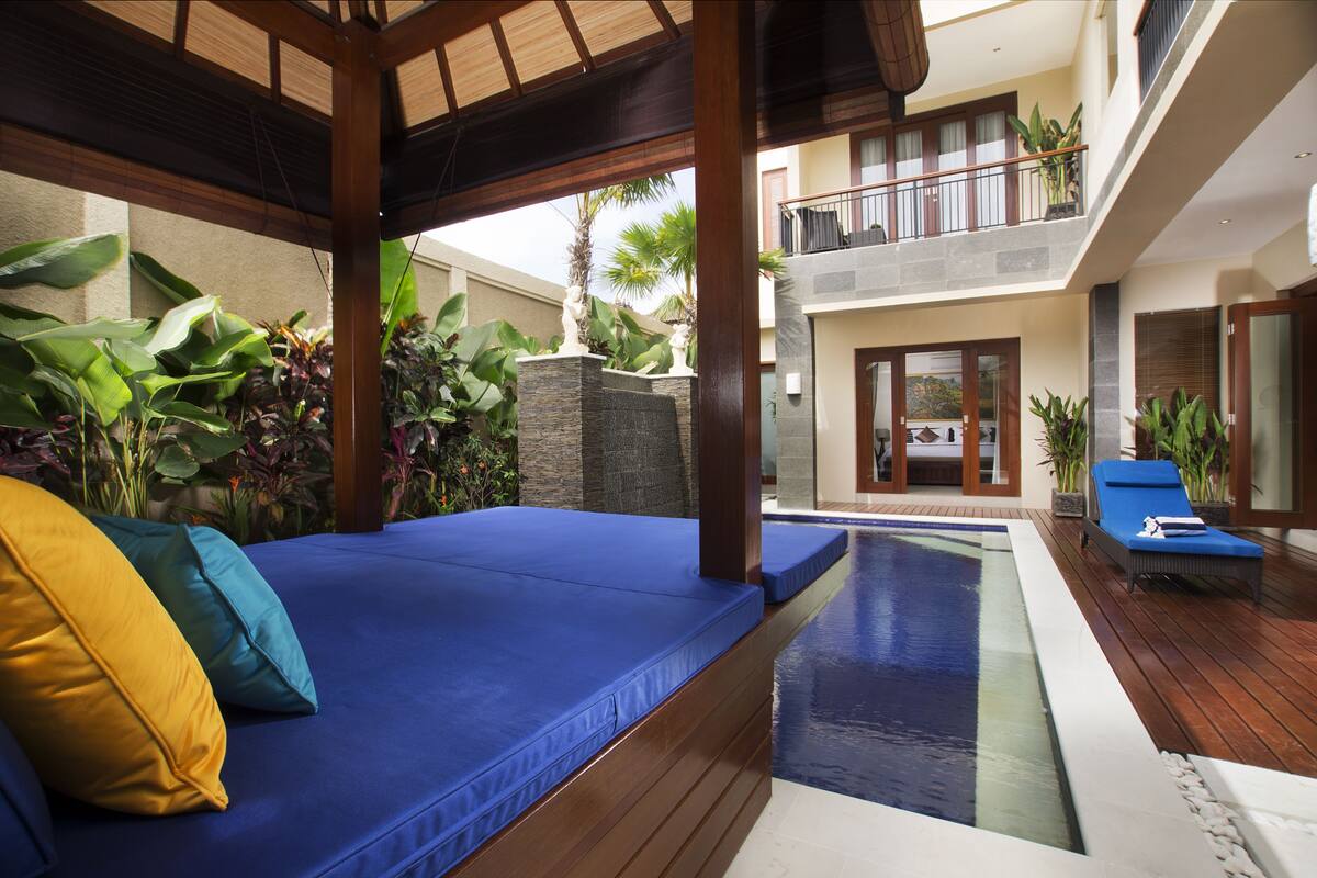 A poolside area with a wooden deck, a blue cushioned seating area, and a view of a building with a balcony.