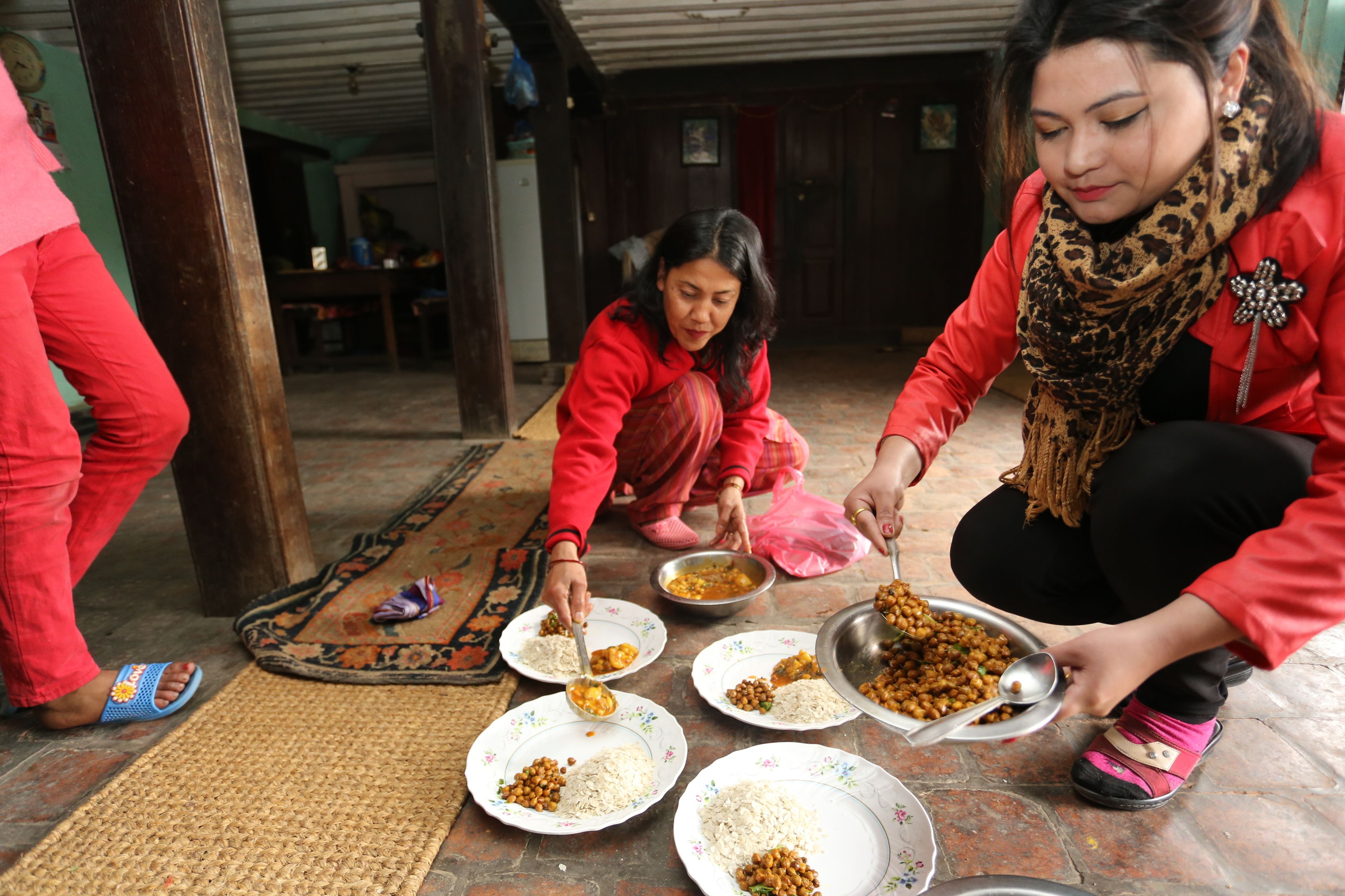 A group of people serving food on the floor in a room with wooden furniture and a patterned mat.