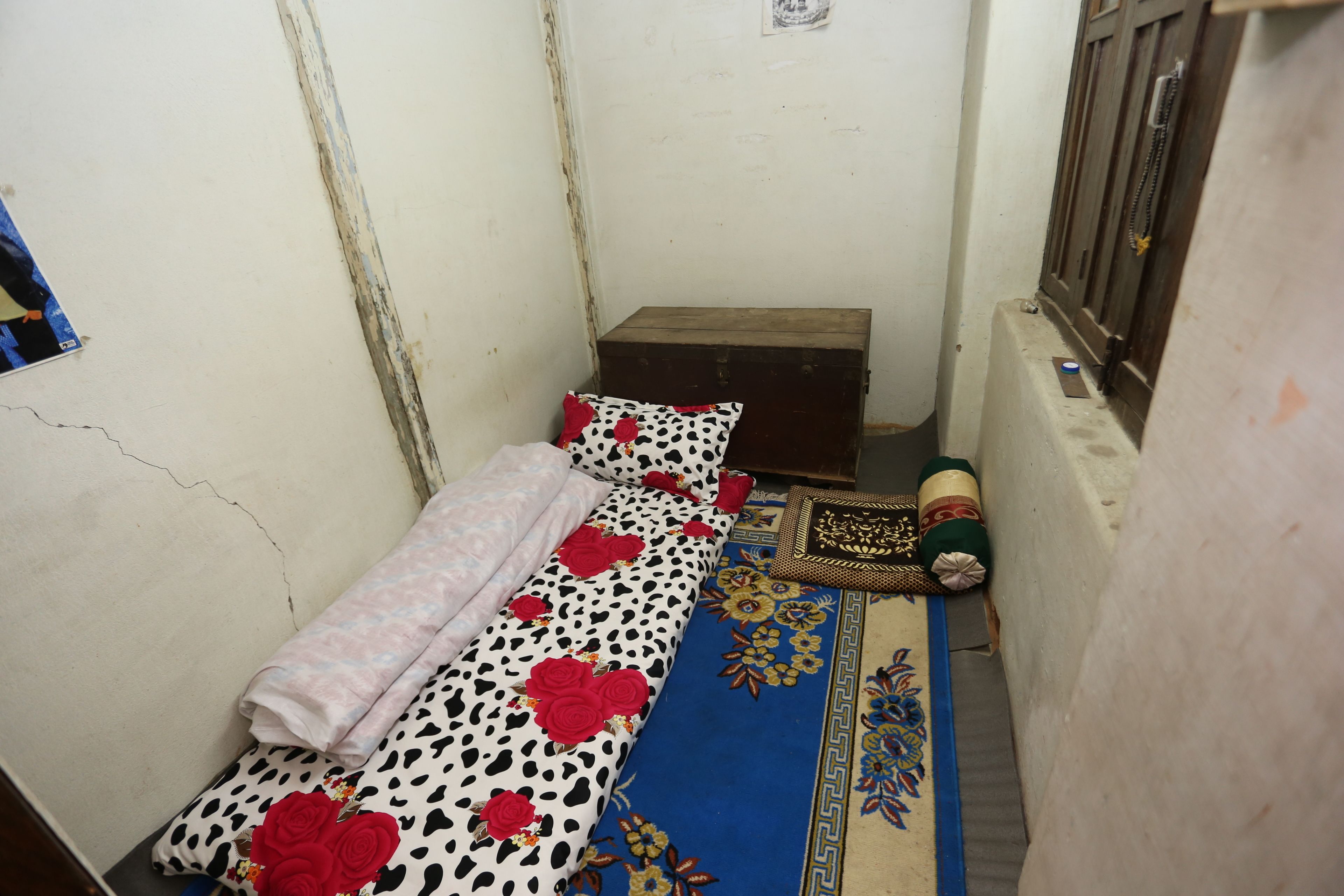 Una habitación pequeña y estrecha con una cama, un cofre de madera y un tapete estampado.