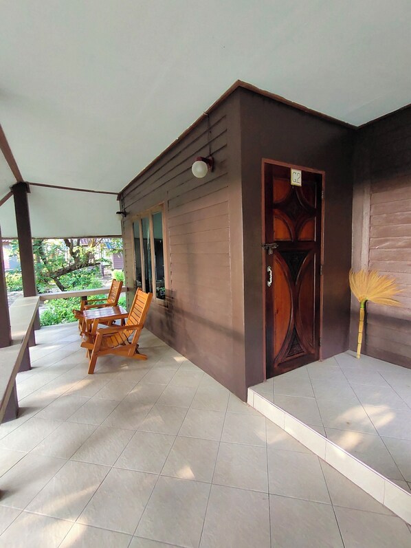 A covered outdoor area with a wooden table and chairs, a door with a sign, and a broom leaning against the wall.