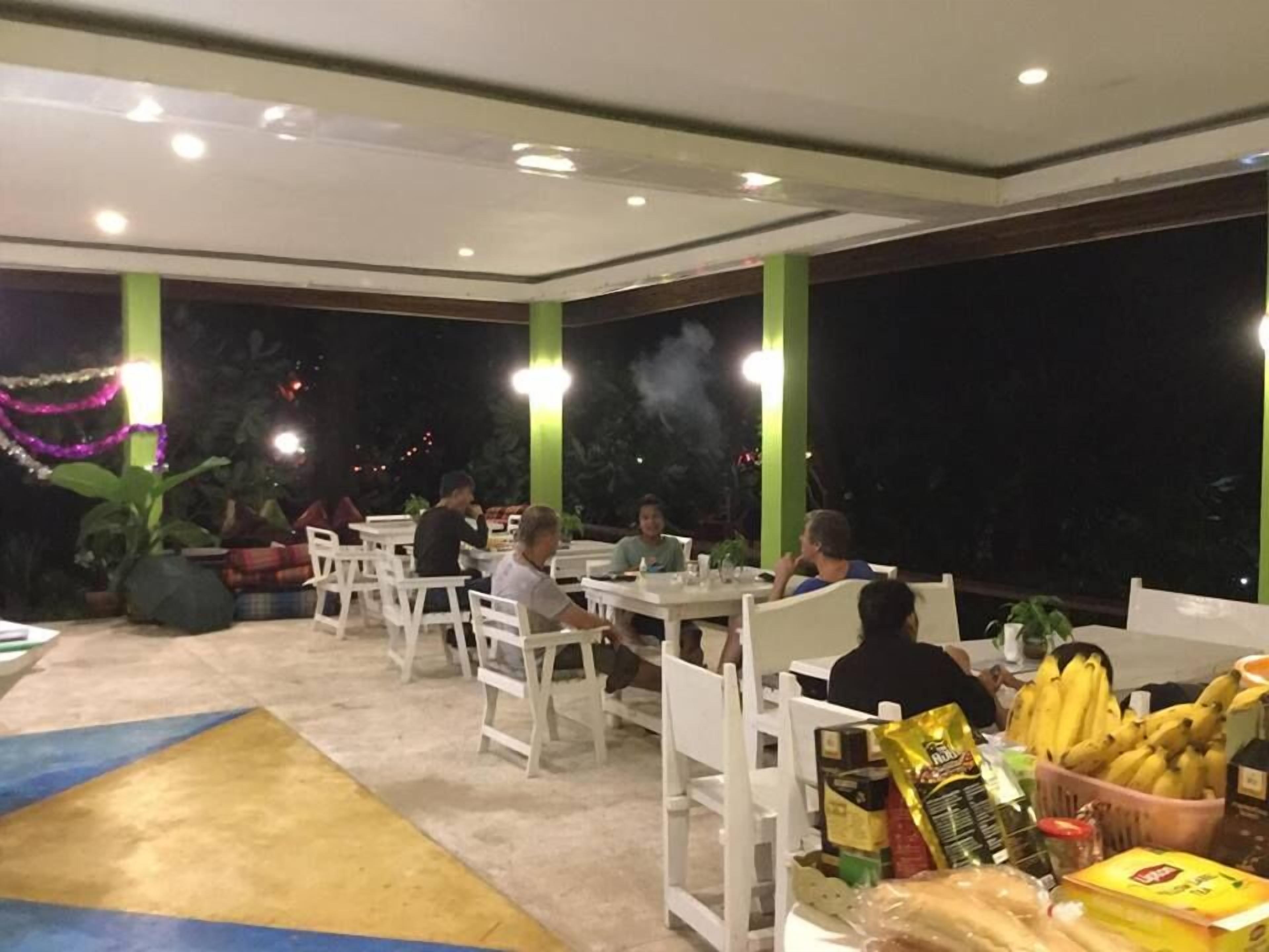 Un área de comedor al aire libre con mesas y sillas blancas, rodeada de follaje y luces decorativas.