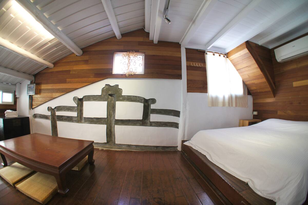 A room with a wooden ceiling, a bed, a table, and a chair.