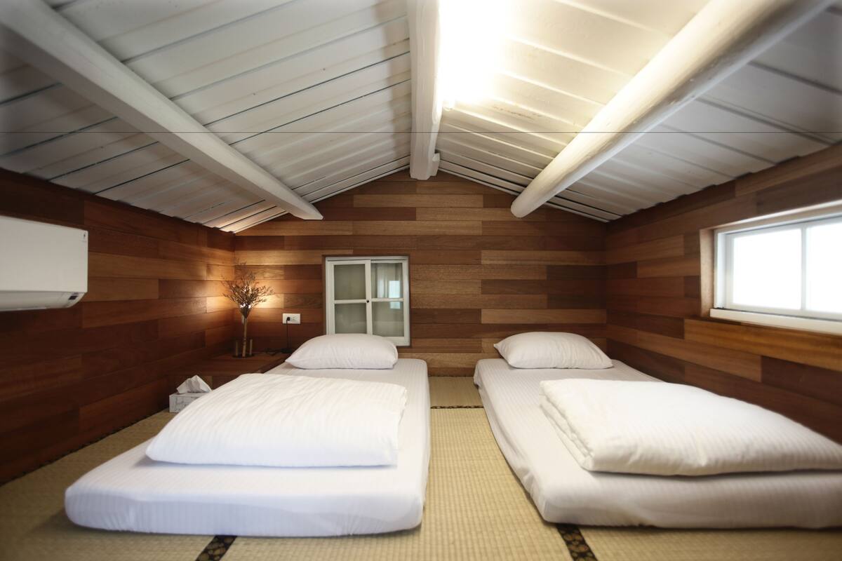 A room with two beds, wooden walls, a window, and a ceiling with exposed beams.
