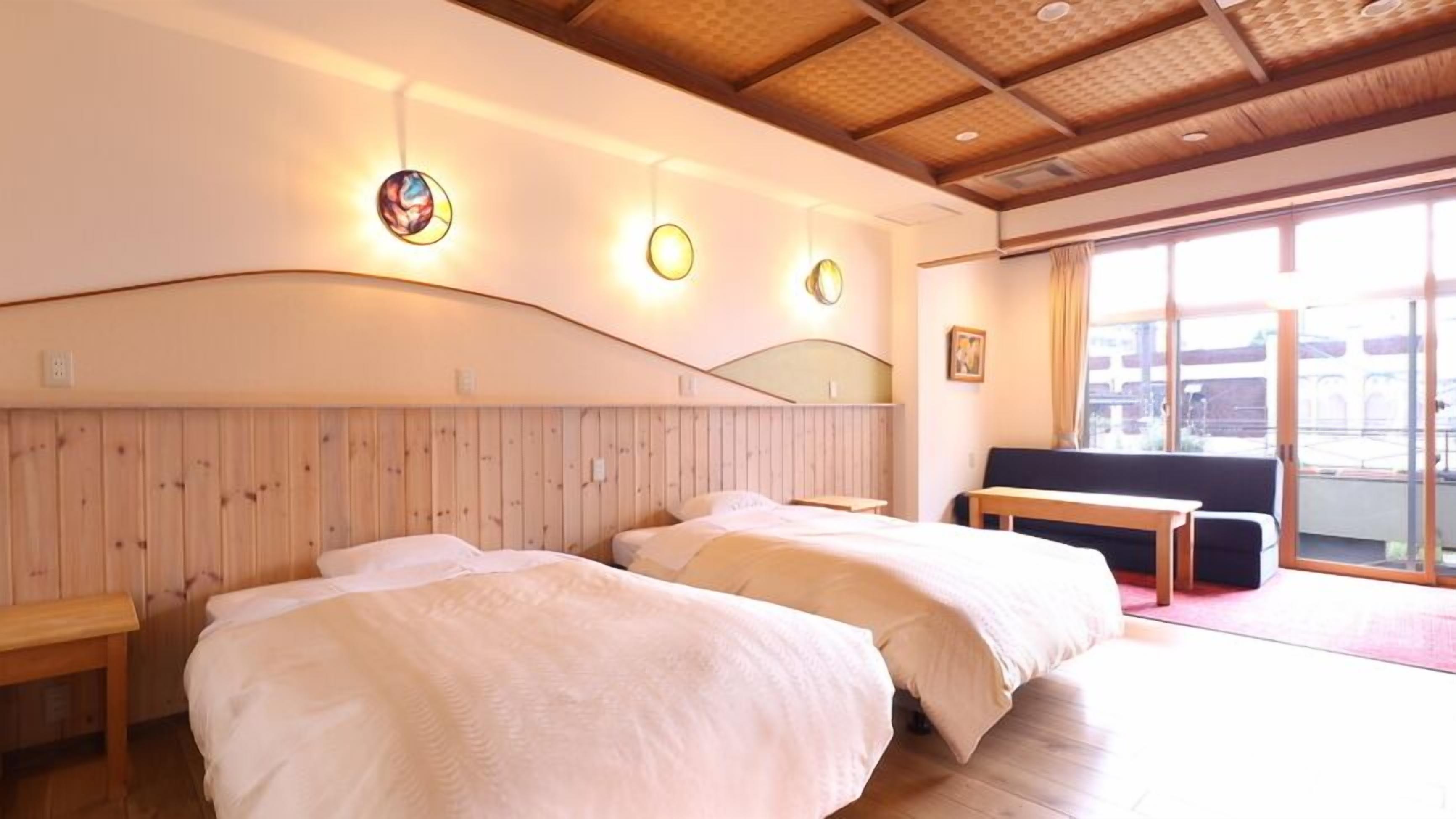 A hotel room with two beds, a wooden headboard, a bench, a large window, and a view of the outside.