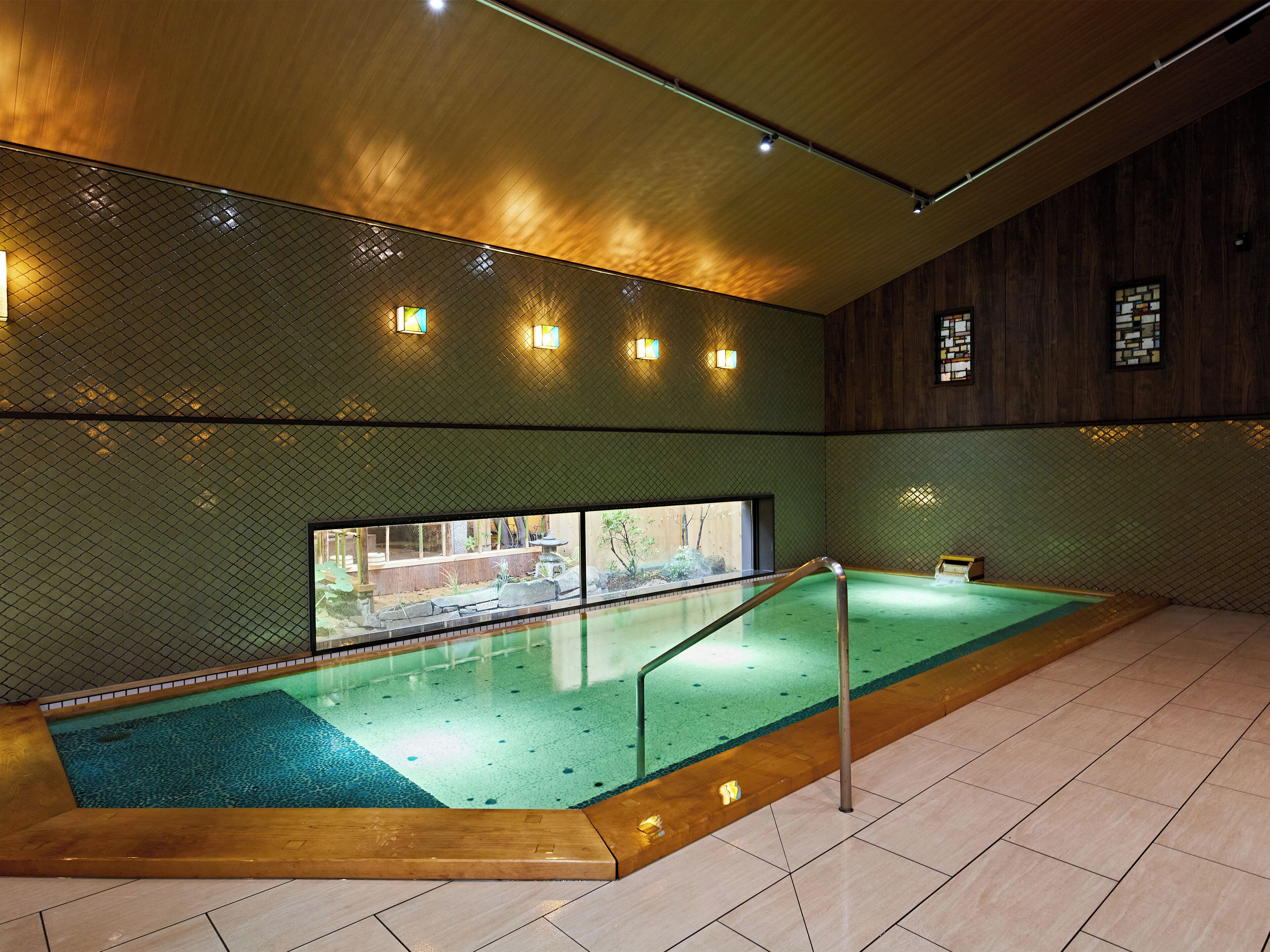 A swimming pool with a wooden deck, a glass wall, and a tiled floor.