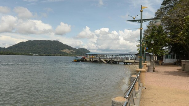 A waterfront area with a dock extending into the water, a pedestrian walkway, and a hill in the background. - Brezza Hotel (Lumut)