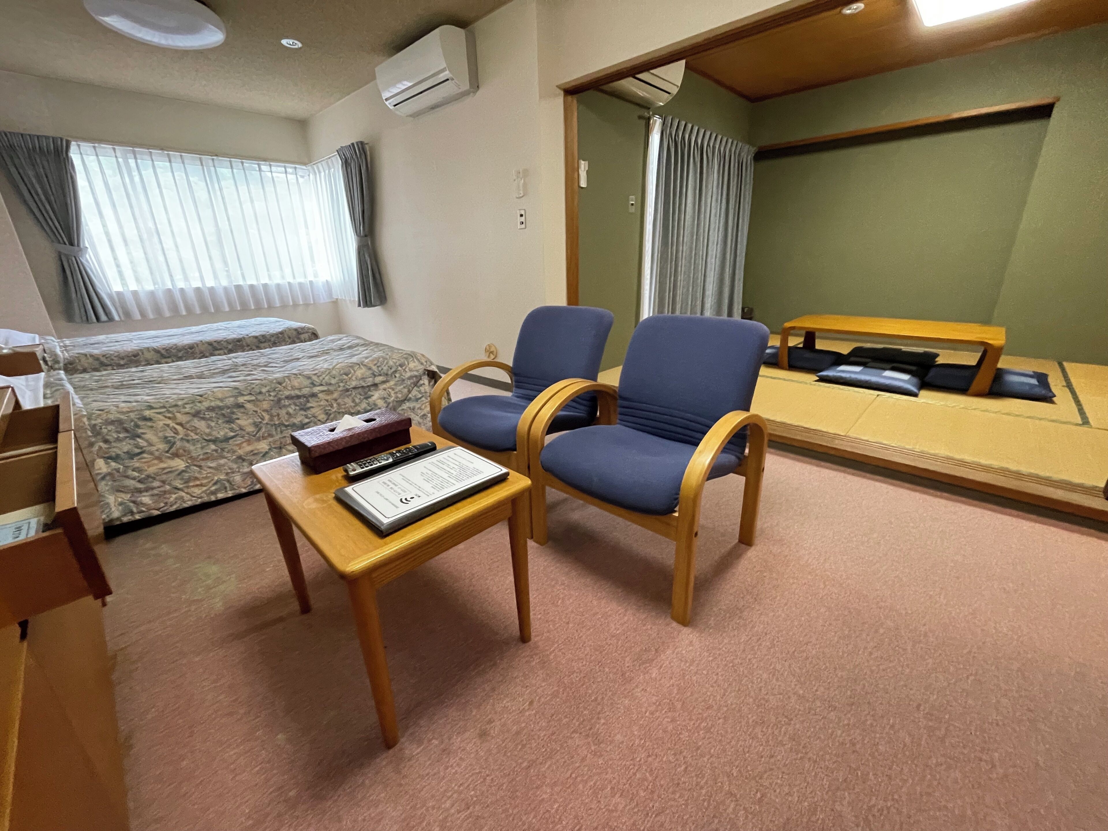 Une chambre d’hôtel avec un lit, deux chaises, une petite table avec une télécommande et une boîte de mouchoirs, et un miroir au mur.
