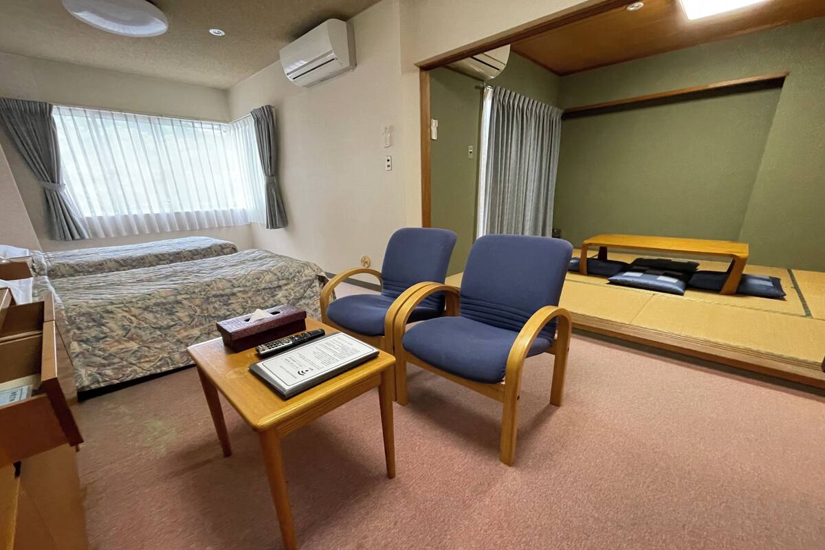 Een hotelkamer met een bed, twee stoelen, een tafeltje met een afstandsbediening en een zakdoos, en een spiegel aan de muur.