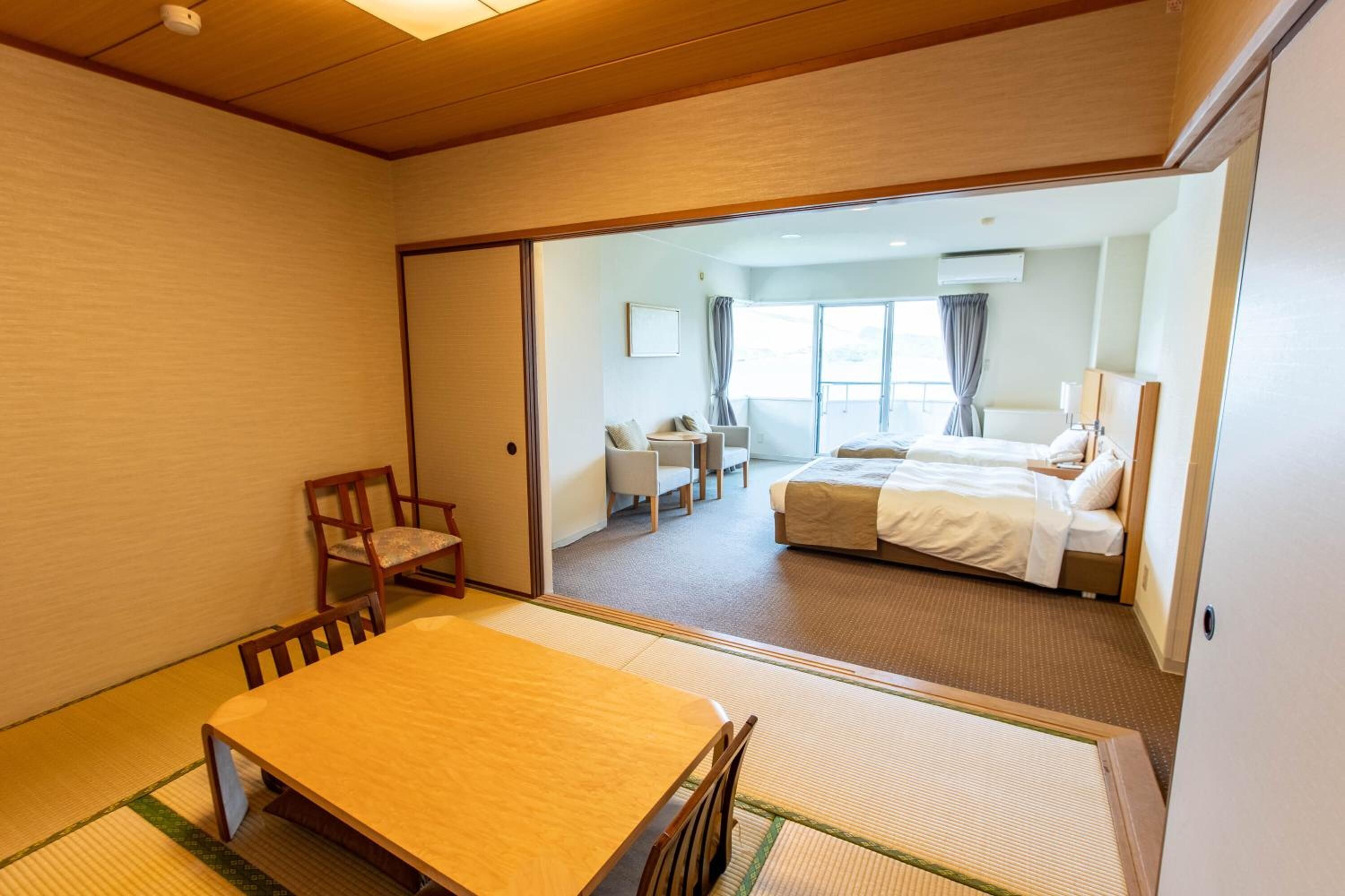 A hotel room with a traditional tatami mat floor, a wooden dining table, a bed with white bedding, and a view of the balcony through a sliding door.