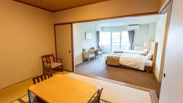 A hotel room with a traditional tatami mat floor, a wooden dining table, a bed with white bedding, and a view of the balcony through a sliding door.