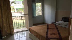 Habitación de hotel con cama, ventana con vista a un edificio verde y palmeras, y balcón con barandilla.