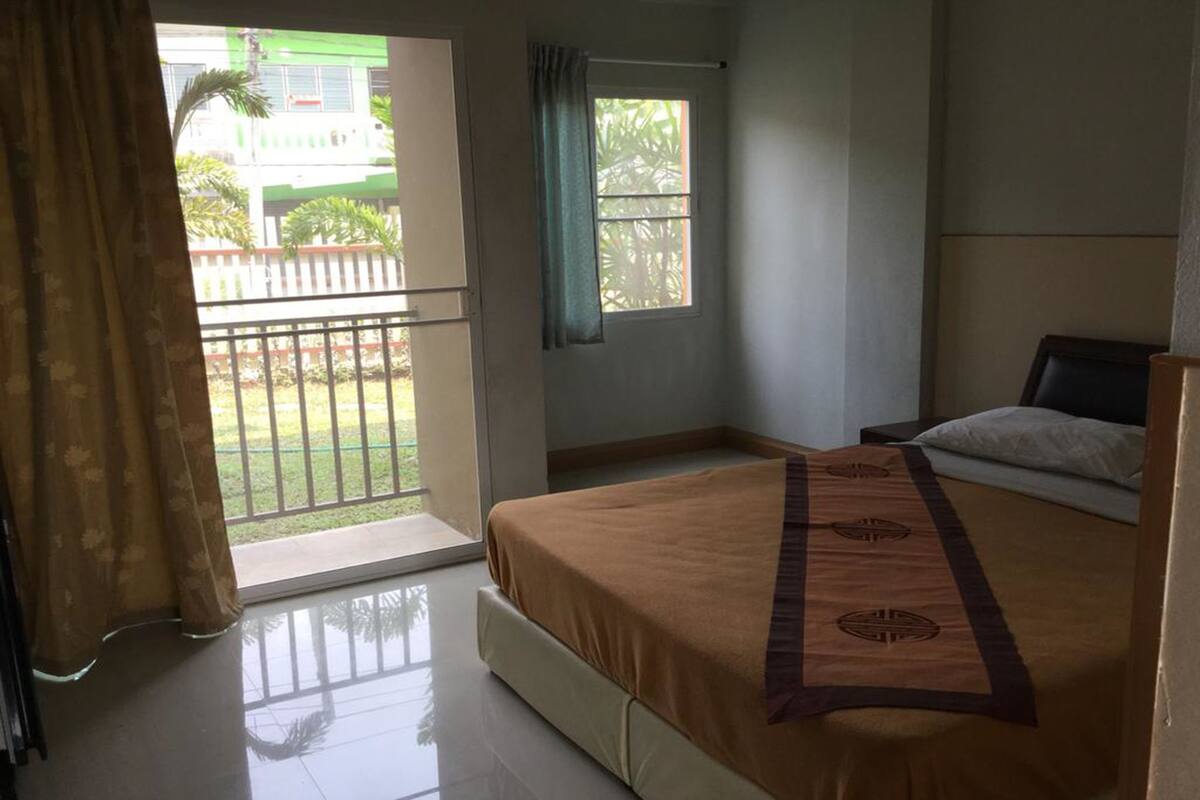 A hotel room with a bed, a window with a view of a green building and palm trees, and a balcony with a railing.