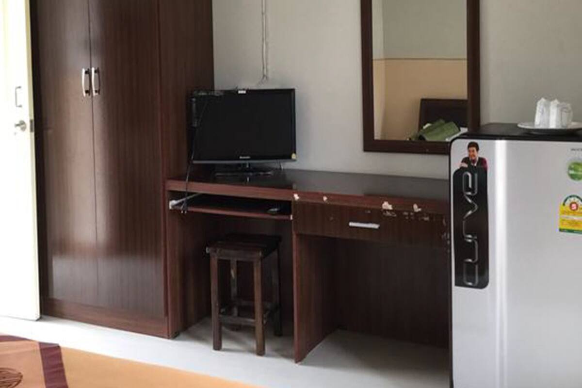 A hotel room with a wooden desk, a television, a refrigerator, a wardrobe, and an air conditioning unit.