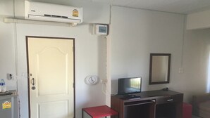 Una habitación con una puerta blanca, un pequeño banco rojo, un escritorio con una computadora, un espejo y un equipo de aire acondicionado.
