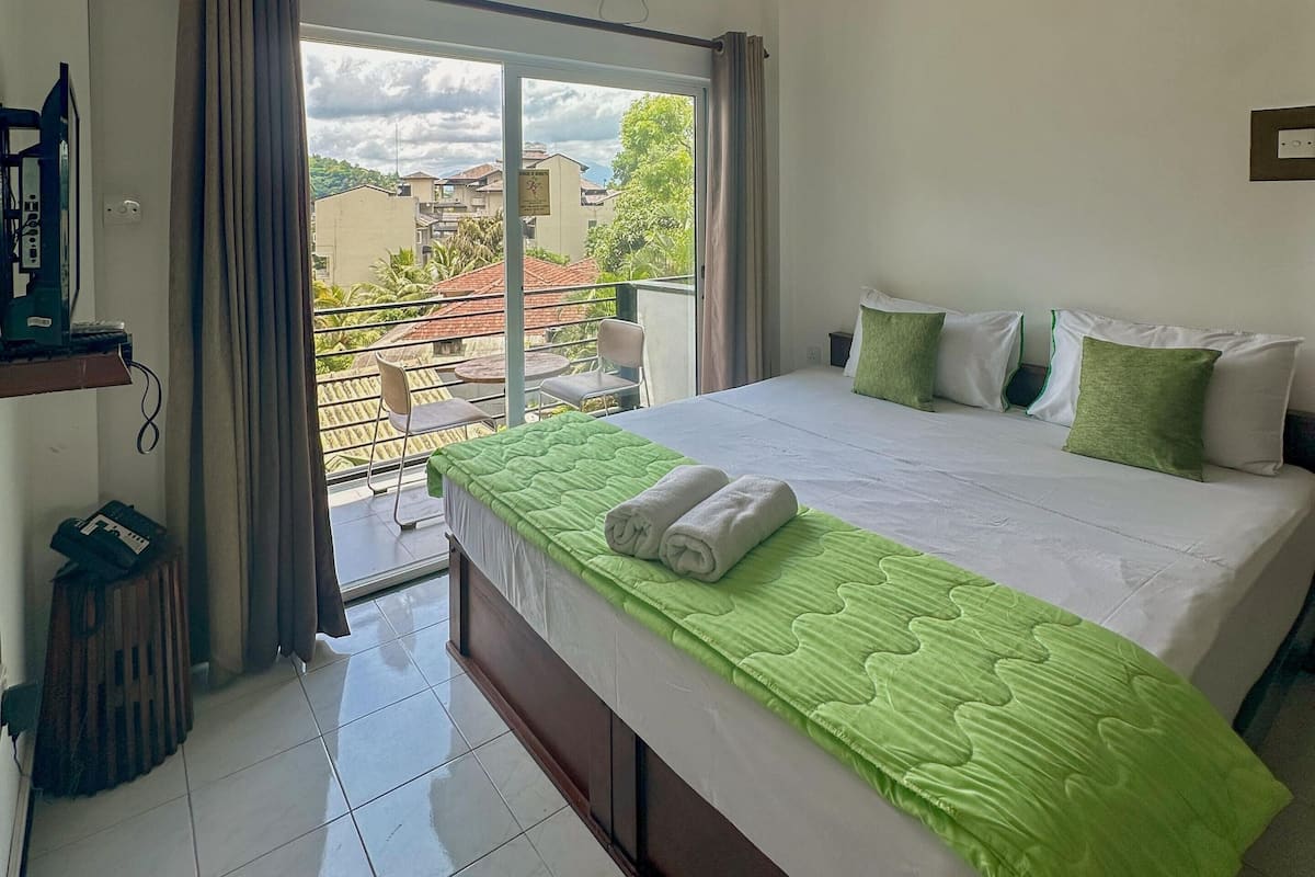 A hotel room with a bed, a small table, a chair, a window with curtains, and an air conditioning unit.
