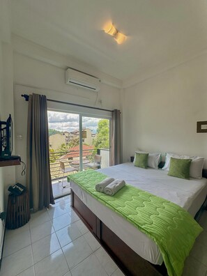 A hotel room with a bed, a small table, a chair, a window with curtains, and an air conditioning unit.