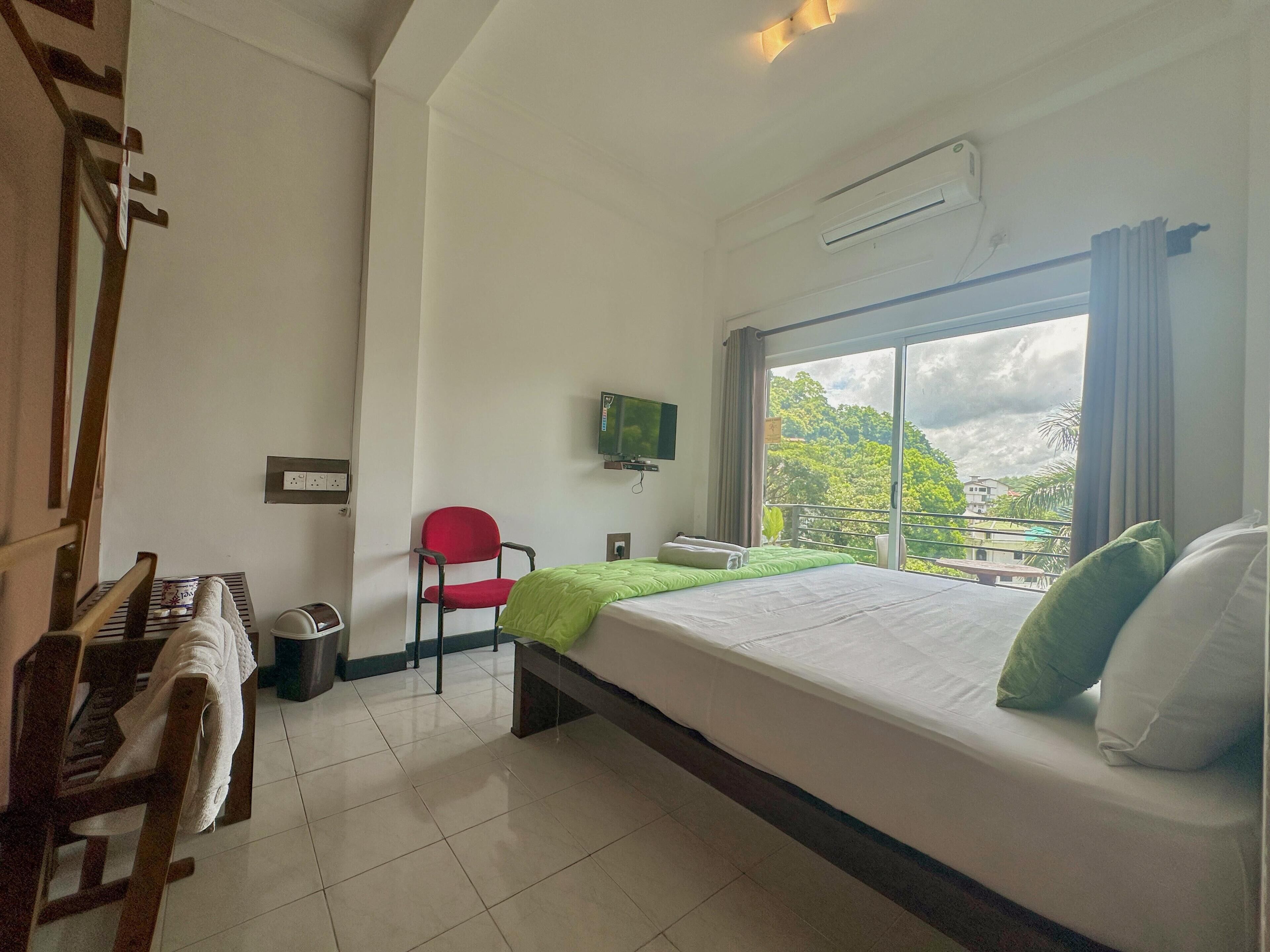 A hotel room with a bed, a TV, a chair, and a window with a view of greenery.