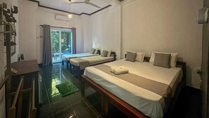 Quarto de hotel com duas camas de solteiro, uma cama grande, ventilador de teto, porta de vidro deslizante para o exterior e uma pequena mesa com cadeira.