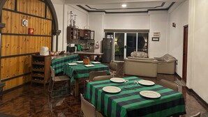 Uma área de refeições com mesas preparadas para quatro pessoas, uma cozinha ao fundo e uma porta de vidro deslizante para uma área externa.