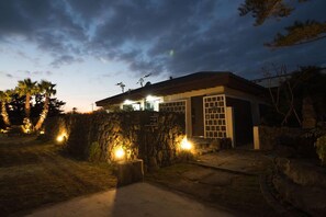A modern house with a stone wall, a garage, and a well-lit driveway at dusk. - Misoga Pension (Seogwipo)