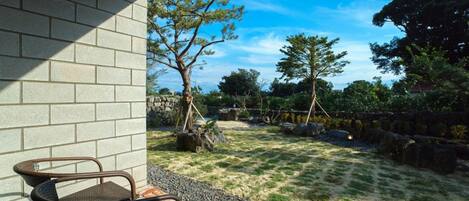 Un patio con muro de ladrillo, una silla de mimbre y un jardín con árboles y un sendero de grava.