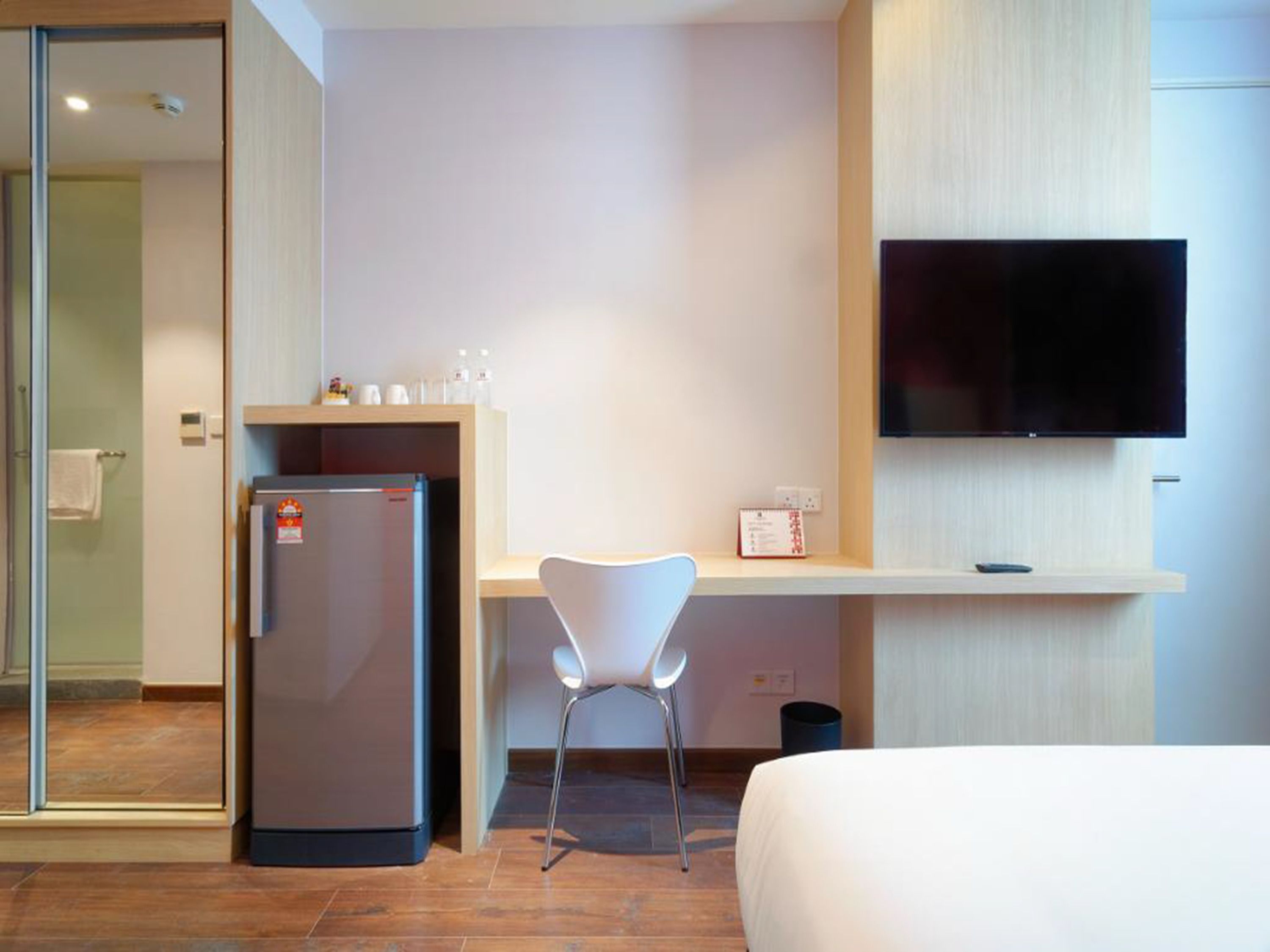 A hotel room with a bed, a desk with a chair, a refrigerator, a TV mounted on the wall, and a small trash bin.