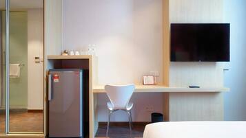 A hotel room with a bed, a desk with a chair, a refrigerator, a TV mounted on the wall, and a small trash bin.