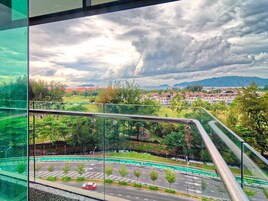 Vista desde un balcón sobre un rotonda con un auto rojo, vegetación y edificios al fondo bajo un cielo nublado.