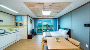 Una habitación de hotel moderna con techo de madera, un ventanal con vistas al mar, una cama con ropa blanca, una mesa de comedor de madera y una zona de cocina con armarios blancos.