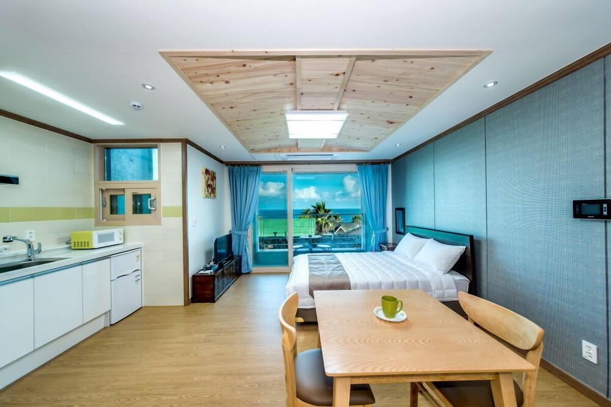 A modern hotel room with a wooden ceiling, a large window with a view of the sea, a bed with white linens, a wooden dining table, and a kitchen area with white cabinets.